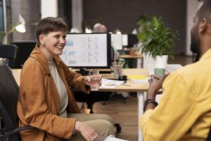 Profissionais conversando em um escritório moderno, com gráficos de desempenho e relatórios digitais em um monitor ao fundo, representando integração de dados e colaboração entre equipes no ambiente corporativo.