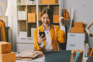 image18840 AnonMedia Empreendedora comemorando uma venda online enquanto segura o celular cercada por caixas de encomendas simbolizando o crescimento do e commerce no 1111 aumento da confiança do consumidor e oportunidades para marcas brasileiras